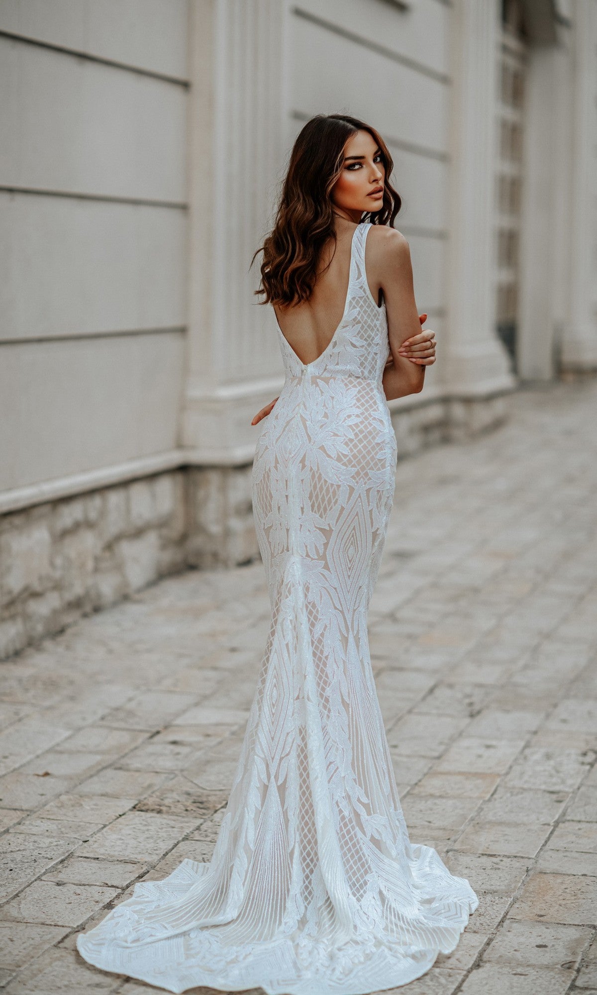 A woman in the Classic Long White Formal Dress: Tina Holly BB052, a sleeveless, form-fitting lace mermaid gown with a low back, stands on a stone walkway and looks over her shoulder.